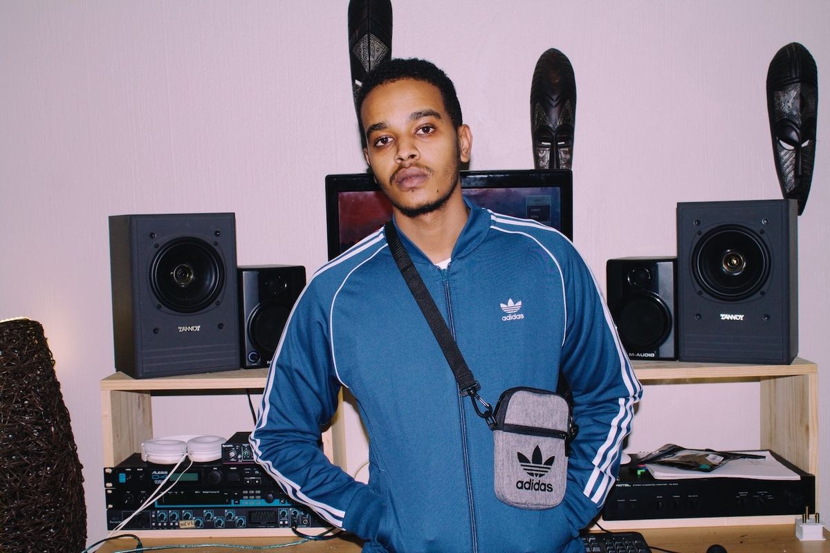 Xplosive DJ posing in his home studio wearing a blue adidas tracksuit and an adidas moon bag. 