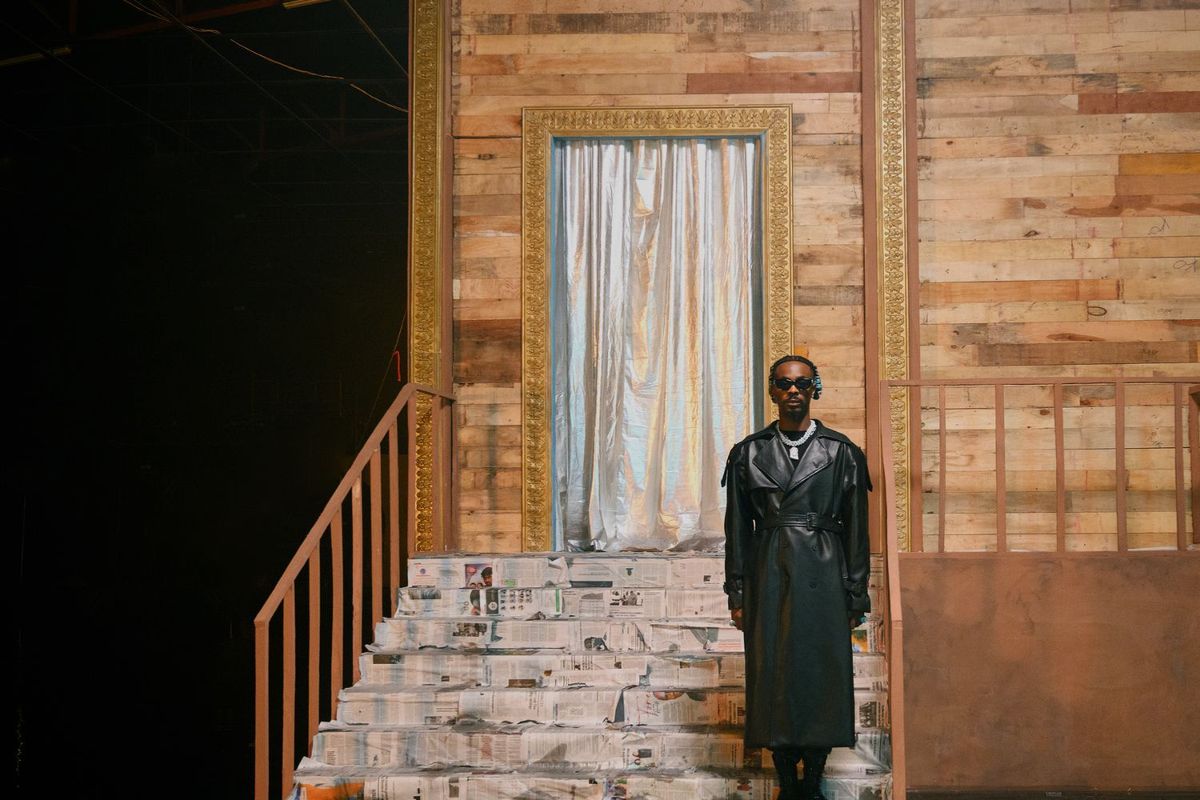 Young Jonn stands in front of the steps of a house, wearing a black leather trench coat, a chunky silver chain, and sunglasses.