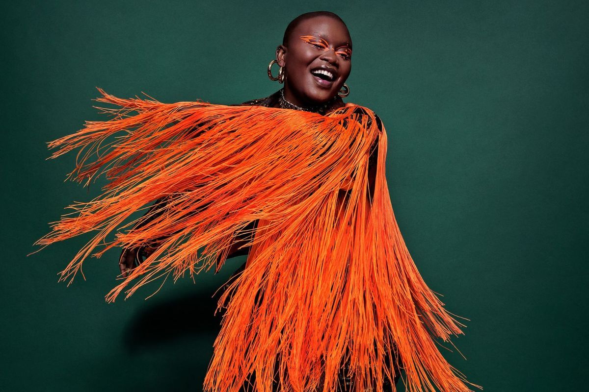 Zoë Modiga takes a moment to laugh while posing for a press picture. Her body is covered in an orange tussel, and orange highlighter decorates her eyelids.